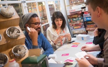 Four adults play cards, with a pile of board games behind them.